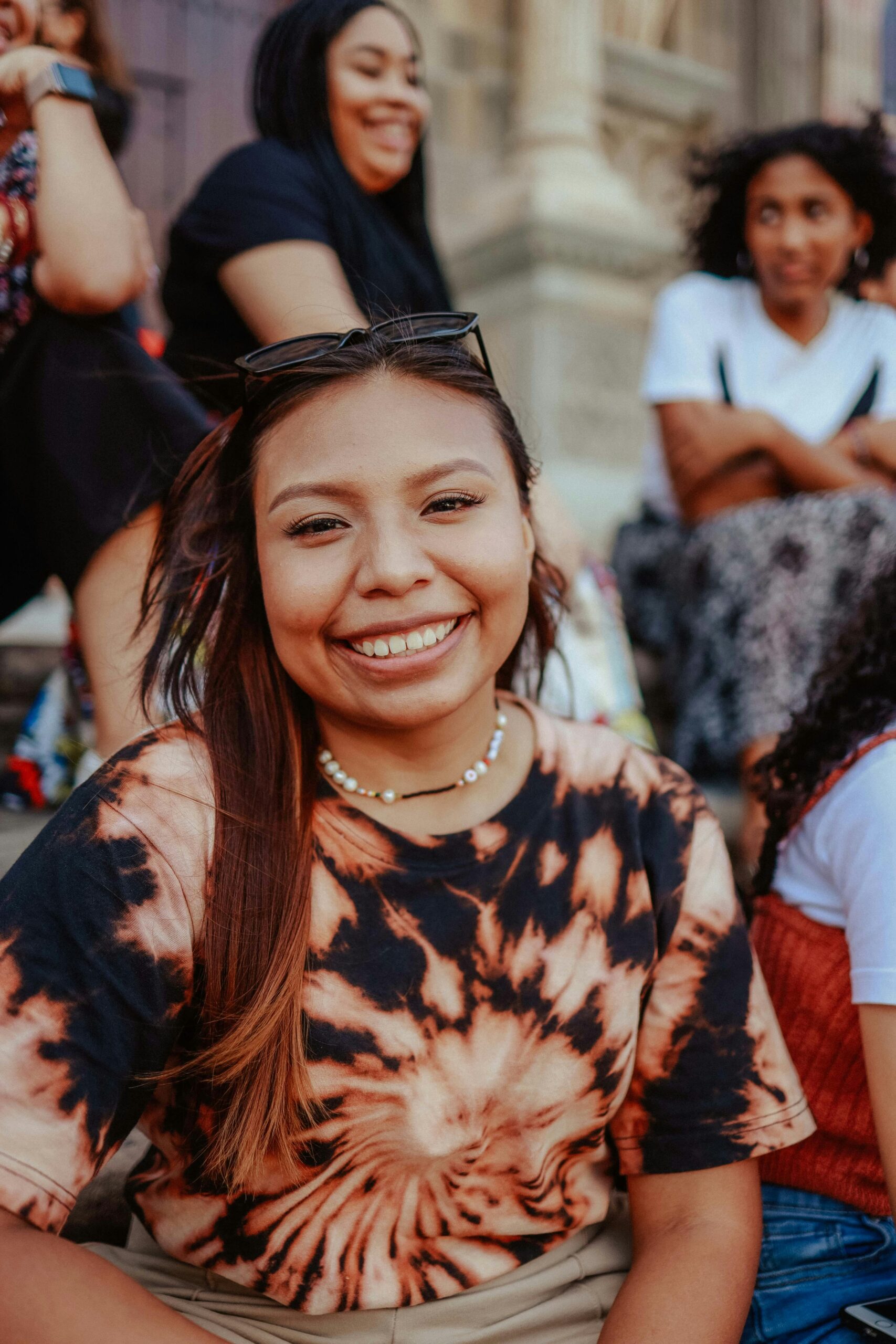A smiling woman with long hair and eyeglasses sits outdoors, wearing a tie-dye shirt. Urban setting.