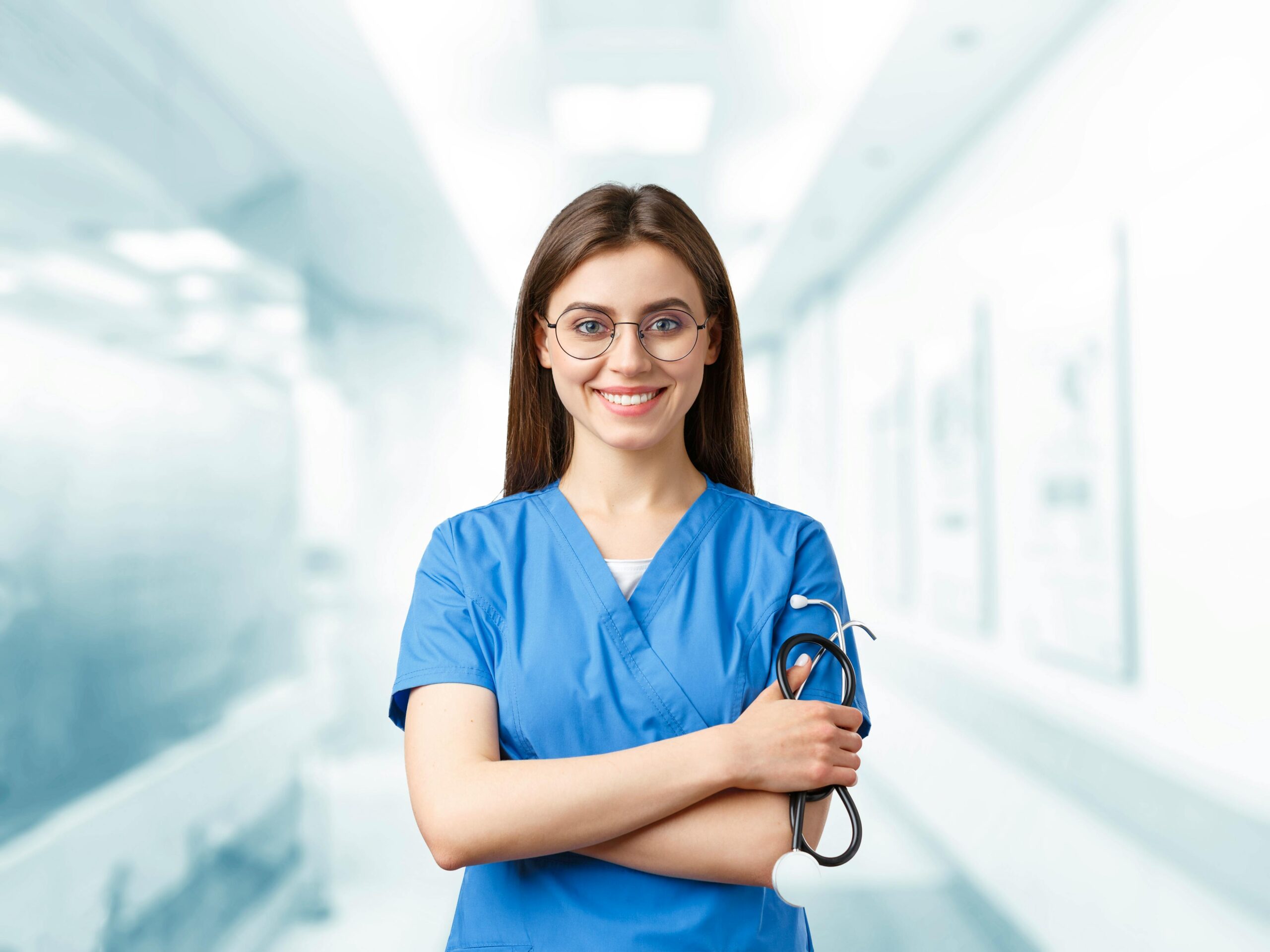 Young nurse in blue scrubs smiling and holding a stethoscope in a hospital corridor.